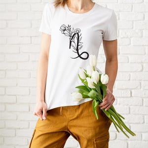 Puede incluir: Una camiseta blanca con un dise&ntilde;o de letra L floral negra. La camiseta la lleva puesta una persona que sostiene un ramo de tulipanes blancos.