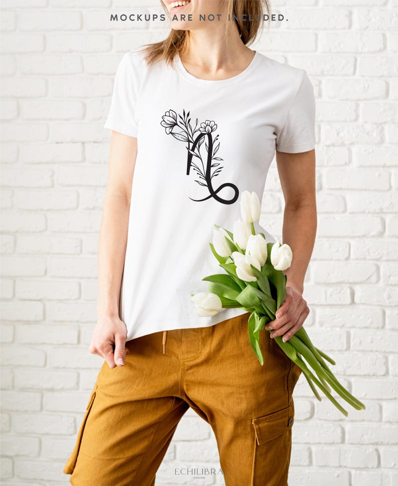 Puede incluir: Una camiseta blanca con un dise&ntilde;o de letra L floral negra. La camiseta la lleva puesta una persona que sostiene un ramo de tulipanes blancos.