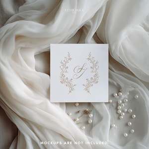 May include: A square white card featuring a gold-colored calligraphic letter S centered between two symmetrical floral branches. The card is displayed against a draped white fabric with scattered white pearls and the text ECHILIBRA.