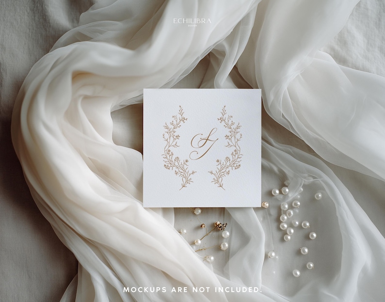 May include: A square white card featuring a gold-colored calligraphic letter S centered between two symmetrical floral branches. The card is displayed against a draped white fabric with scattered white pearls and the text ECHILIBRA.