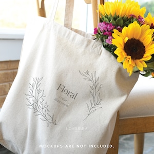 May include: A beige tote bag with a floral illustration and the word "Floral". The bag is filled with bright yellow sunflowers and small purple flowers. The bag is resting on a wooden chair.