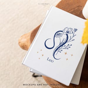 Puede incluir: Un diario blanco con un dise&ntilde;o floral azul y dorado que representa el signo del zod&iacute;aco Leo. El diario tiene el texto "Leo" impreso en la portada.