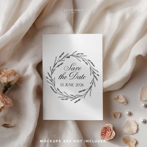 May include: A white card with the text "Save the Date" inside a floral wreath. The date "15 JUNE 2026" is below the text. The card is on a cream-colored surface with roses, petals, and pearls.