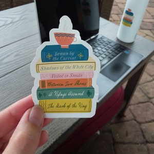 May include: A sticker depicting a stack of books, including titles such as "Drawn by the Current" and "Shadows of the White City." A teacup with steam sits on top. The sticker is held in a hand, with a laptop and water bottle visible.