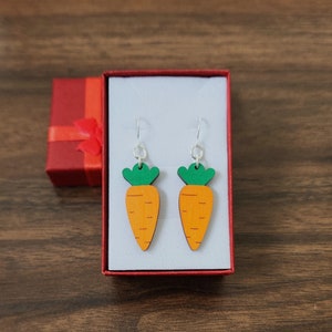 May include: A pair of carrot-shaped earrings in a red and white gift box. The earrings are made of wood and painted orange with green tops.