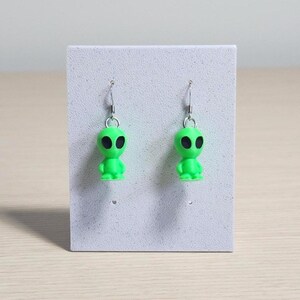 May include: A pair of lime green alien earrings with black eyes and silver-colored ear wires. The earrings are displayed on a white card, set against a light wood-grain surface.