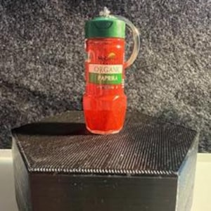 May include: A clear glass jar with a red lid and a green ring around the lid. The jar is filled with red paprika and the label reads "Organic Paprika".