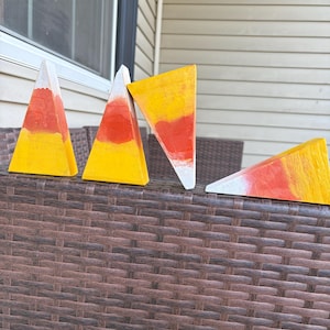 May include: Four triangular wooden blocks painted in a candy corn pattern with horizontal bands of white, orange, and yellow. Three blocks stand upright in a vertical orientation, while one block is positioned horizontally on its side.