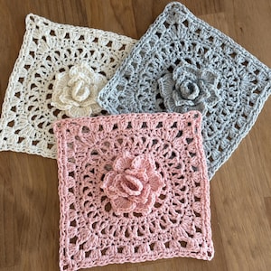 Puede incluir: Tres cuadrados de ganchillo hechos a mano en crema, gris y rosa, cada uno con un diseño floral central. Los cuadrados están dispuestos sobre una superficie de madera, mostrando los intrincados detalles del ganchillo.