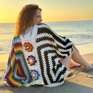 May include: A woman wearing a colorful crochet top with a black and white striped pattern. The top features a rainbow design and floral accents. The woman is sitting on a sandy beach with the ocean in the background.
