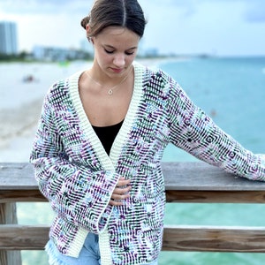 May include: A woman wearing a multi-coloured crocheted cardigan with a white trim. The cardigan has a geometric pattern with shades of purple, green, and white. She is wearing denim shorts and a black vest top.
