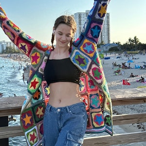 Op de afbeelding: Een vrouw draagt een kleurrijk gehaakt vest met een sterpatroon. Het vest is gemaakt van verschillende gekleurde vierkanten met sterren in het midden. De vrouw draagt een blauwe jeans en een zwart tanktop.