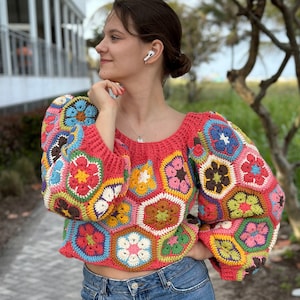 Może przedstawiać: Kolorowy top na szydełku z geometrycznym wzorem kwiatów w odcieniach różowego, zielonego, żółtego, brązowego i białego. Top ma luźny krój i długie rękawy.