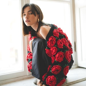 May include: A woman wearing a black top with a red crocheted rose cardigan. The cardigan is made of many red roses and green leaves.