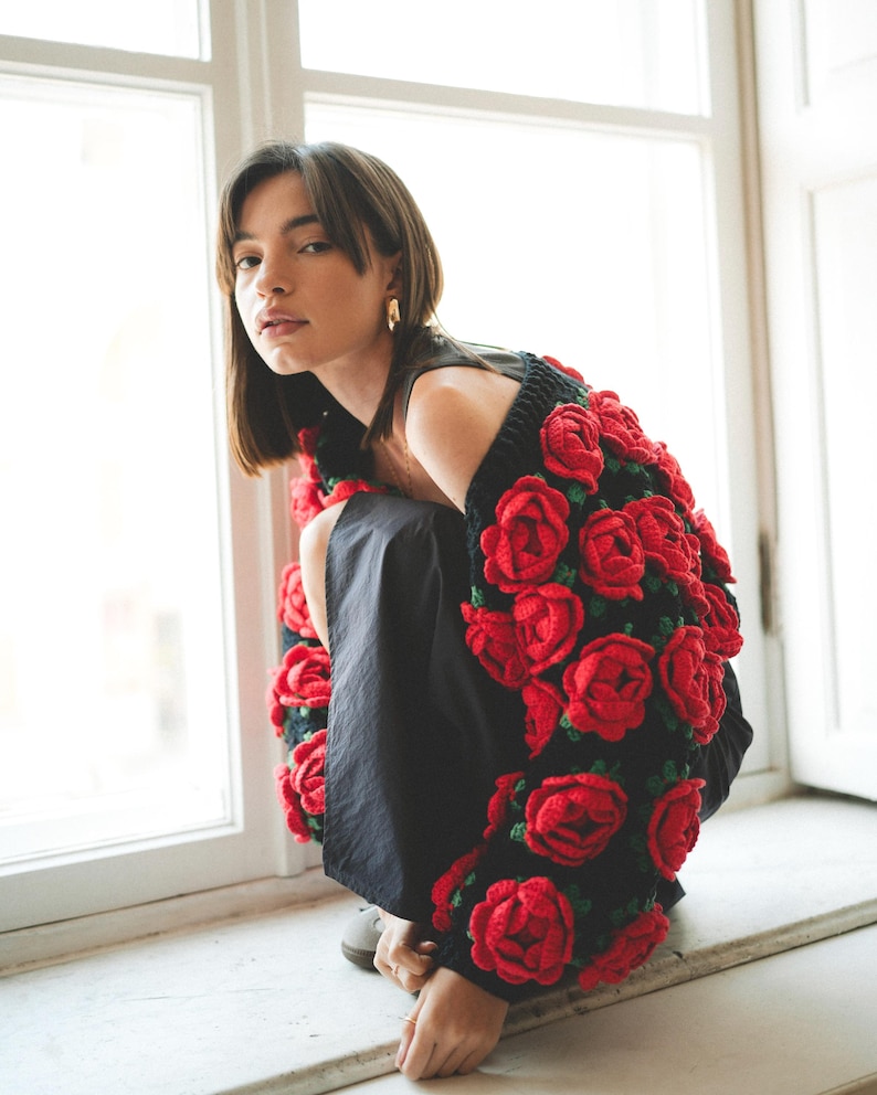 May include: A woman wearing a black top with a red crocheted rose cardigan. The cardigan is made of many red roses and green leaves.