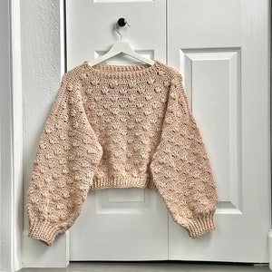May include: A beige crocheted sweater with a textured, bobble stitch pattern. The sweater has a relaxed fit and long sleeves.