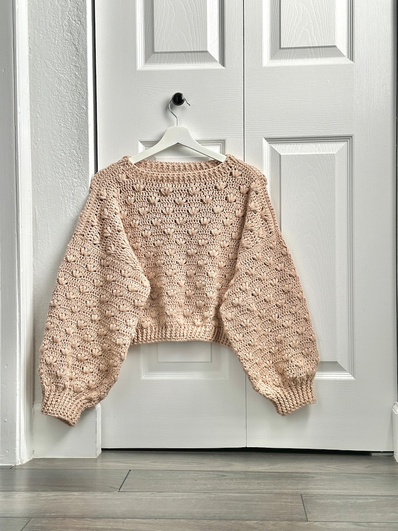 May include: A beige crocheted sweater with a textured, bobble stitch pattern. The sweater has a relaxed fit and long sleeves.