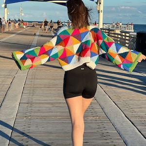 May include: A person wearing a colorful crocheted sweater with a geometric pattern. The sweater has a cropped style and is made with a variety of bright colors, including pink, blue, green, and yellow. The person is walking on a wooden pier.