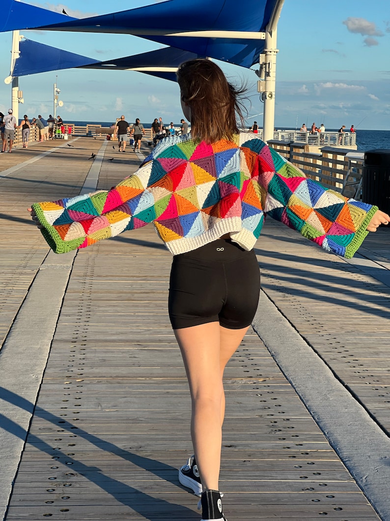 May include: A person wearing a colorful crocheted sweater with a geometric pattern. The sweater has a cropped style and is made with a variety of bright colors, including pink, blue, green, and yellow. The person is walking on a wooden pier.