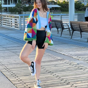 May include: A woman wearing a colorful crocheted cardigan with a geometric pattern, a white tank top, black shorts, and black high-top sneakers. She is standing on a wooden pier.