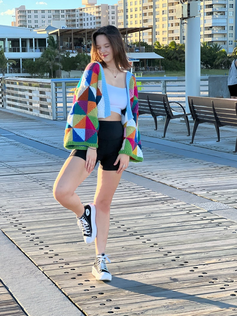May include: A woman wearing a colorful crocheted cardigan with a geometric pattern, a white tank top, black shorts, and black high-top sneakers. She is standing on a wooden pier.