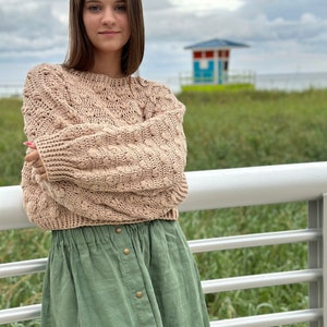 May include: A woman wearing a light brown knitted sweater with a textured pattern and a green button-down skirt.