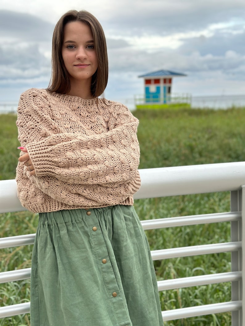May include: A woman wearing a light brown knitted sweater with a textured pattern and a green button-down skirt.