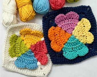 May include: Two crocheted flower squares in white and navy blue, with colorful petals in shades of green, yellow, orange, pink, and blue. Several skeins of yarn in various colors are arranged above the squares, including white, yellow, blue, and pink.