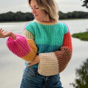 May include: A long-sleeve knit jumper featuring a colour-blocked design with horizontal sections of turquoise, blue, pink, orange, beige, and brown. The garment has a ribbed crew neckline and wide, voluminous sleeves with a fuzzy, textured yarn finish.