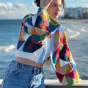 May include: A colorful crocheted cardigan with a patchwork design. The cardigan is cropped and has long sleeves. The person wearing the cardigan is standing in front of a body of water.