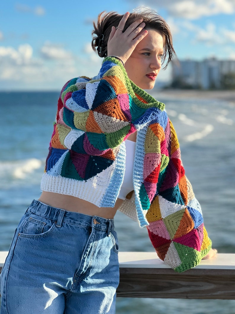 May include: A colorful crocheted cardigan with a patchwork design. The cardigan is cropped and has long sleeves. The person wearing the cardigan is standing in front of a body of water.