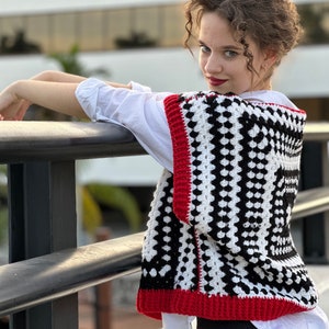May include: A black and white crochet vest with red trim. The vest has a square pattern and is worn over a white shirt.