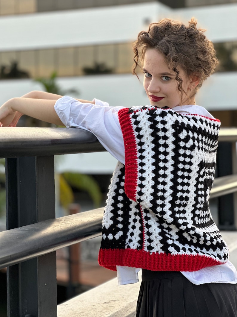 May include: A black and white crochet vest with red trim. The vest has a square pattern and is worn over a white shirt.