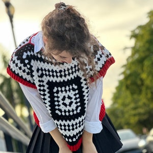 May include: A black and white crochet top with red trim. The top has a geometric pattern and is worn over a white shirt. The person is wearing a black skirt and black tights.