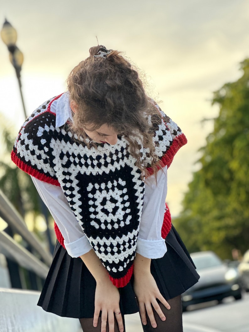 May include: A black and white crochet top with red trim. The top has a geometric pattern and is worn over a white shirt. The person is wearing a black skirt and black tights.