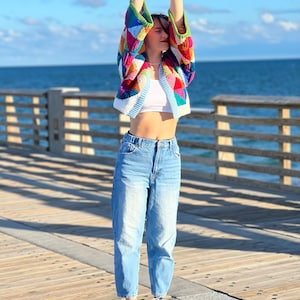 May include: A woman wearing a colorful crocheted cardigan with a white tank top and blue jeans. She is standing on a wooden pier with the ocean in the background.