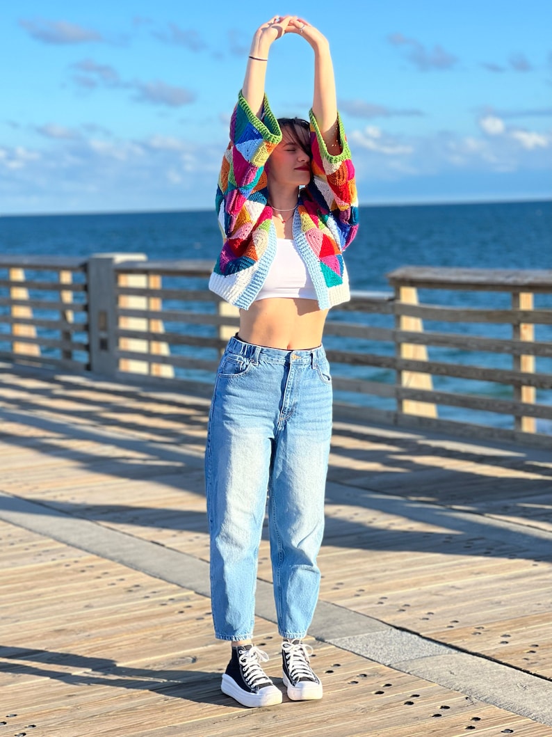 May include: A woman wearing a colorful crocheted cardigan with a white tank top and blue jeans. She is standing on a wooden pier with the ocean in the background.