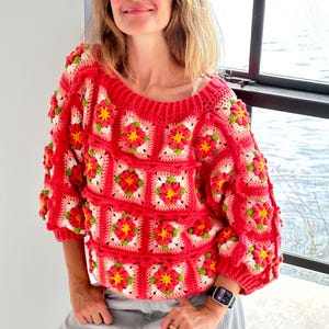 May include: A red crochet sweater with a floral pattern. The sweater features a square design with red, white, yellow, and green flowers. The sleeves are three-quarter length. The person is wearing gray pants.