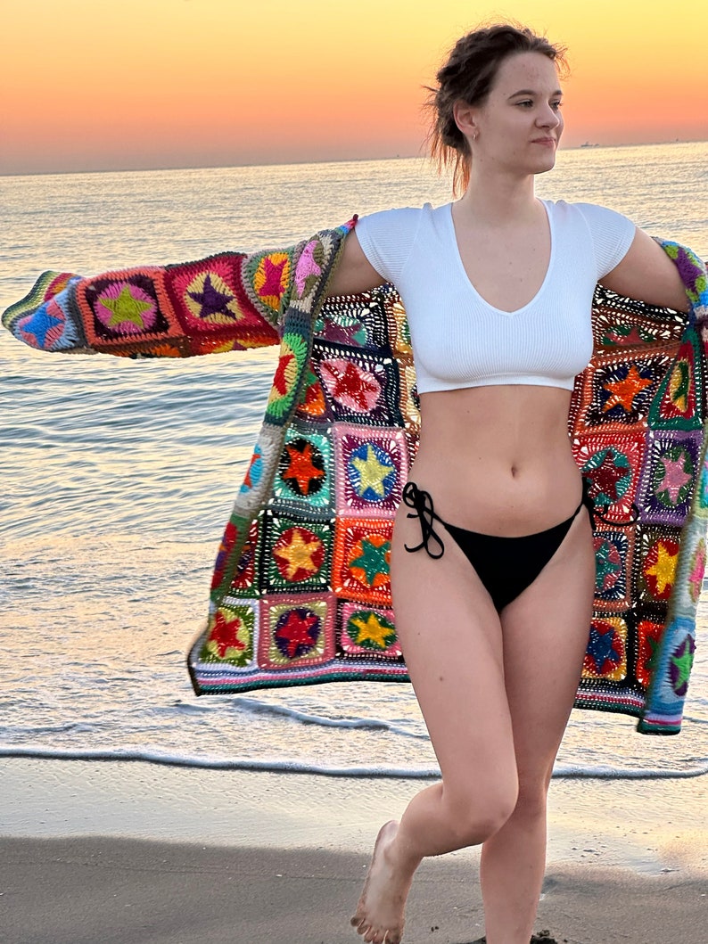Op de afbeelding: Een vrouw draagt een kleurrijk gehaakt vest met een sterpatroon over een wit crop top en een zwarte bikini broek. Ze staat op een zandstrand met de oceaan op de achtergrond.