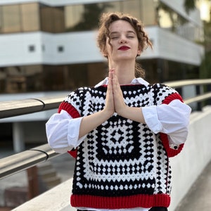 May include: A woman wearing a white shirt with a black and white crochet vest with red trim. The vest has a square pattern.
