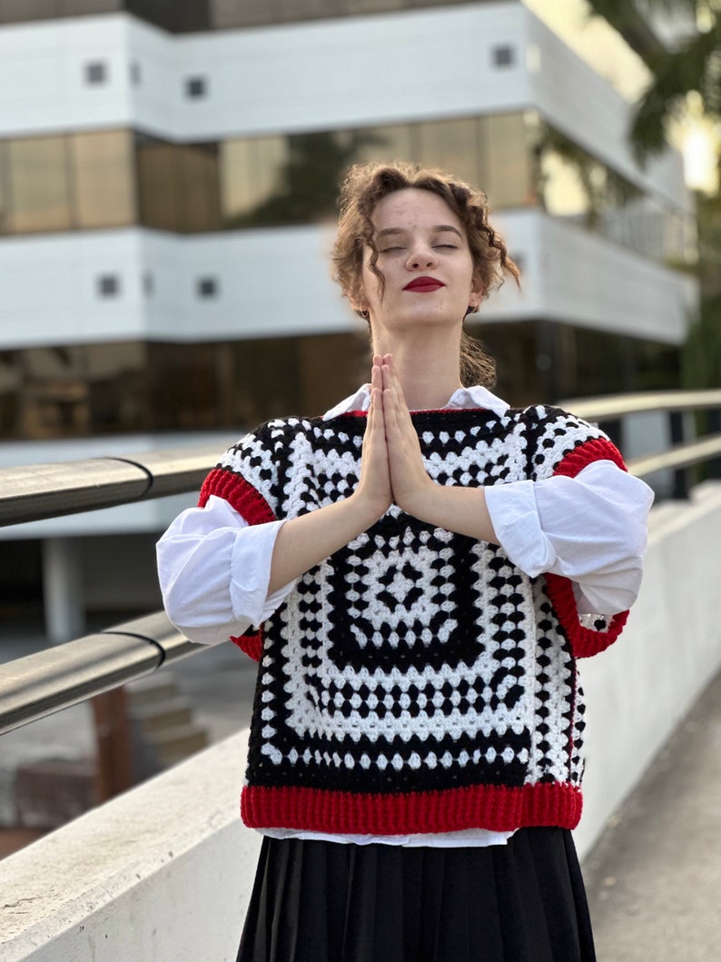 May include: A woman wearing a white shirt with a black and white crochet vest with red trim. The vest has a square pattern.