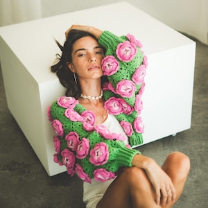 May include: A woman wearing a green and pink crocheted cardigan with large flower embellishments. The cardigan is cropped and has a loose fit.