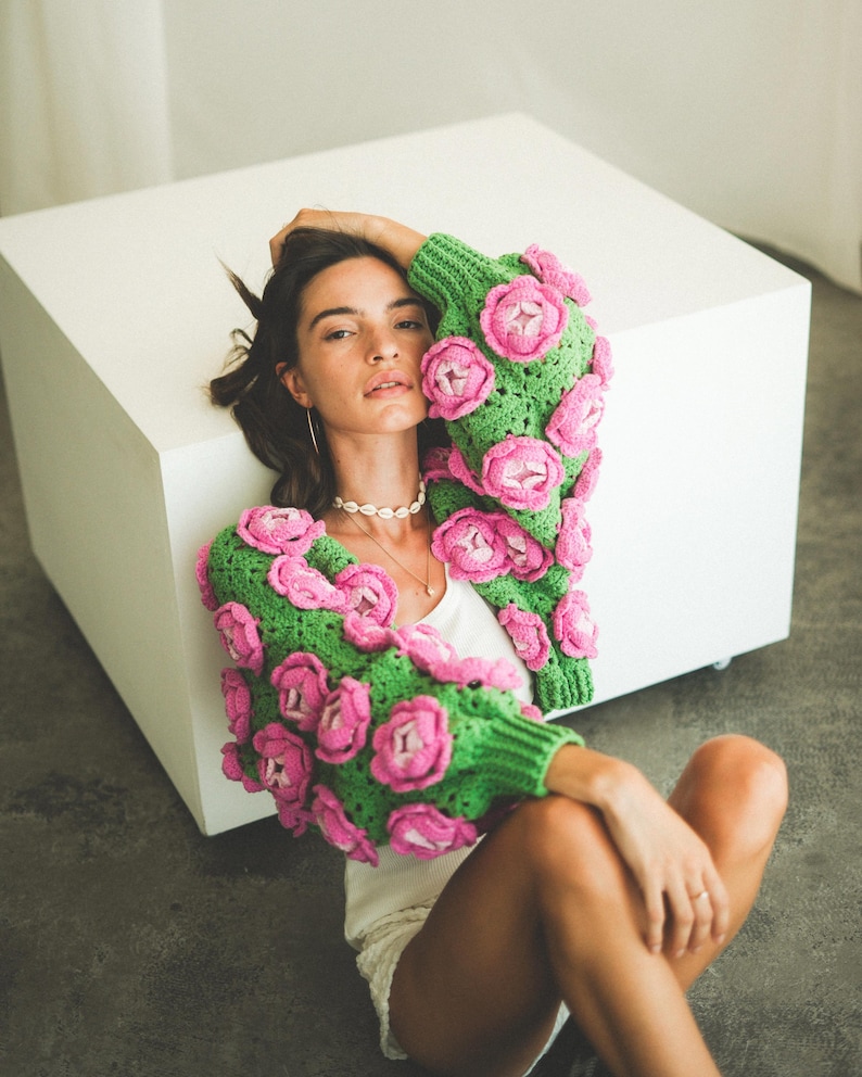 May include: A woman wearing a green and pink crocheted cardigan with large flower embellishments. The cardigan is cropped and has a loose fit.