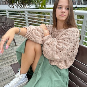 May include: A woman wearing a light pink knitted sweater and a green skirt sits on a wooden bench. She is wearing white high-top sneakers with a black star logo.