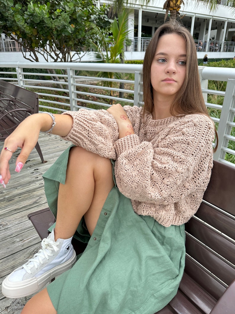 May include: A woman wearing a light pink knitted sweater and a green skirt sits on a wooden bench. She is wearing white high-top sneakers with a black star logo.