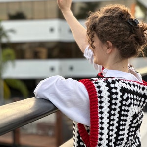 May include: A woman wearing a white shirt and a black and white crocheted vest with red trim. The vest has a geometric pattern.