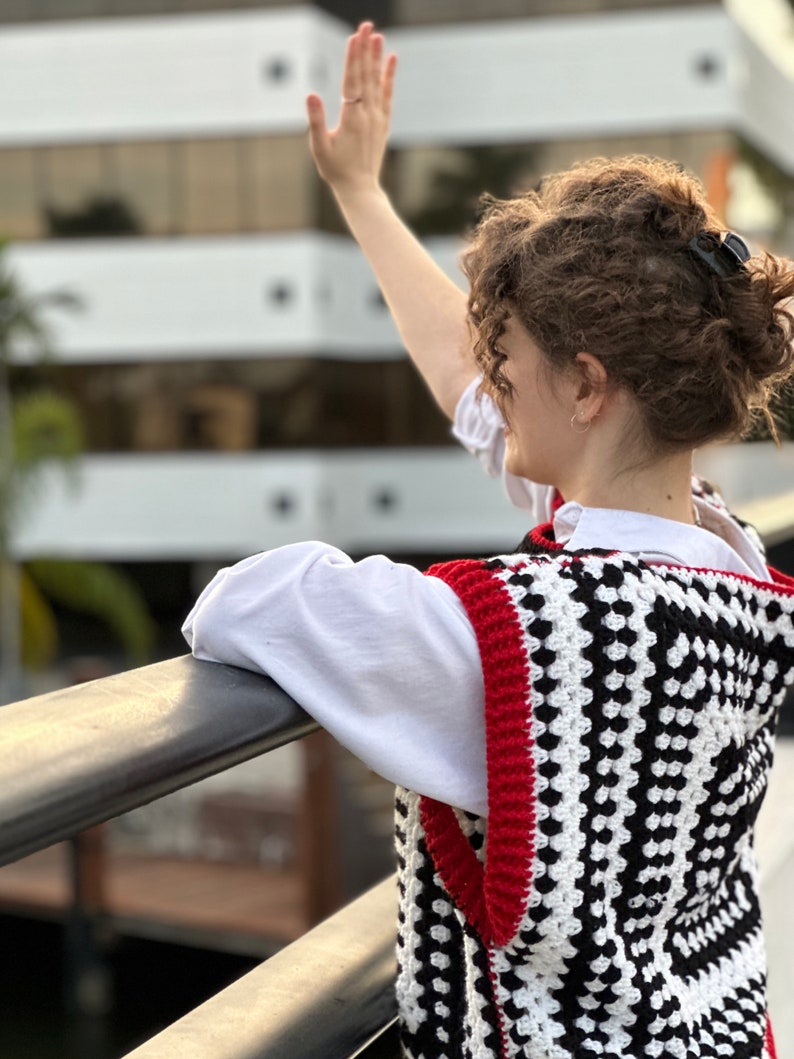 May include: A woman wearing a white shirt and a black and white crocheted vest with red trim. The vest has a geometric pattern.