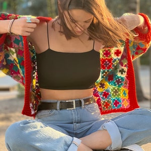 May include: A woman wearing a colorful crochet cardigan over a black tank top, blue jeans, and black sneakers.