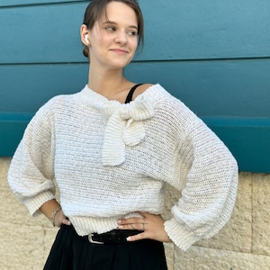 Könnte beinhalten: Ein weißer Häkelpullover mit einem Schleifendetail am Ausschnitt. Der Pullover hat eine lockere Passform und lange Ärmel.