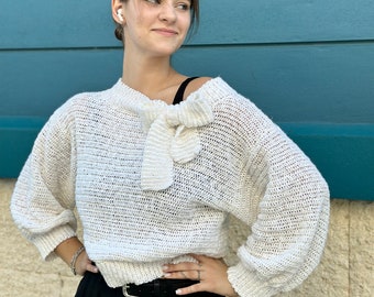 May include: A white crocheted sweater with a bow detail at the neckline. The sweater has a loose fit and long sleeves.